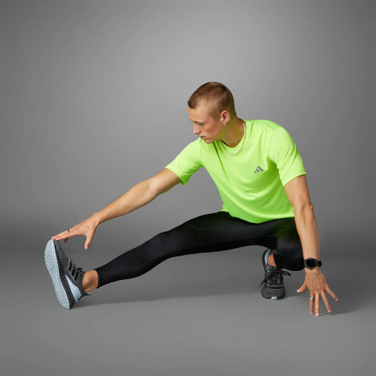 A man wearing a t-shirt and black adidas running tights stretches his leg out to the side while wearing running shoes.