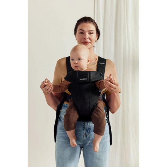 Woman holding a baby in a black and brown baby carrier against a white background