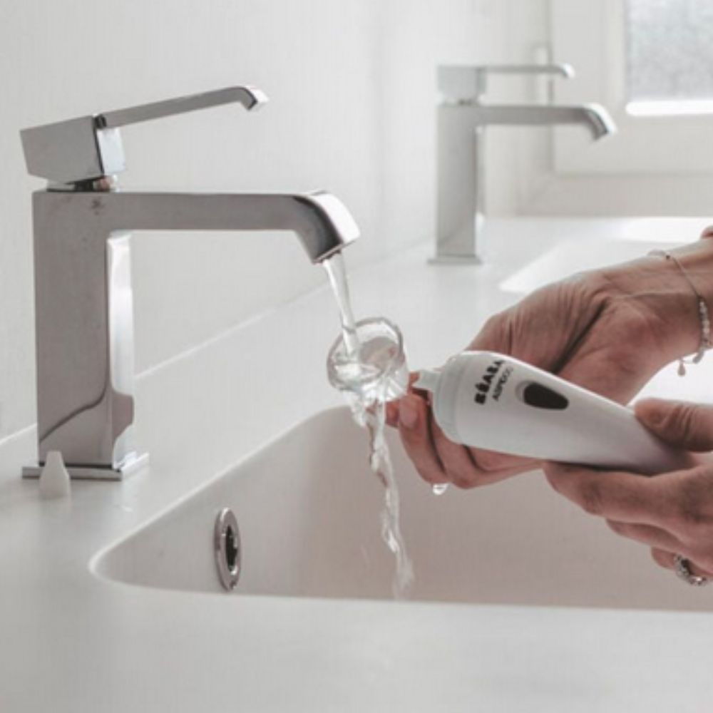 Person washing Beaba Aspidoo Nasal Aspirator under water flow from a faucet in a bathroom setting.