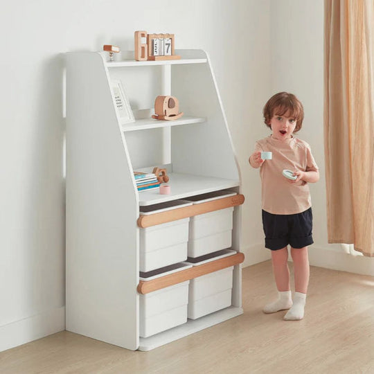 Child standing next to a white bookshelf with storage bins in a room with a curtain.