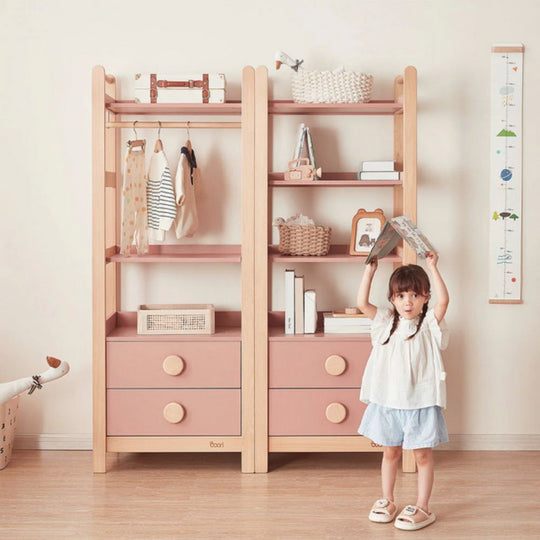Child standing next to a wooden and pink children's wardrobe with shelves and drawers.