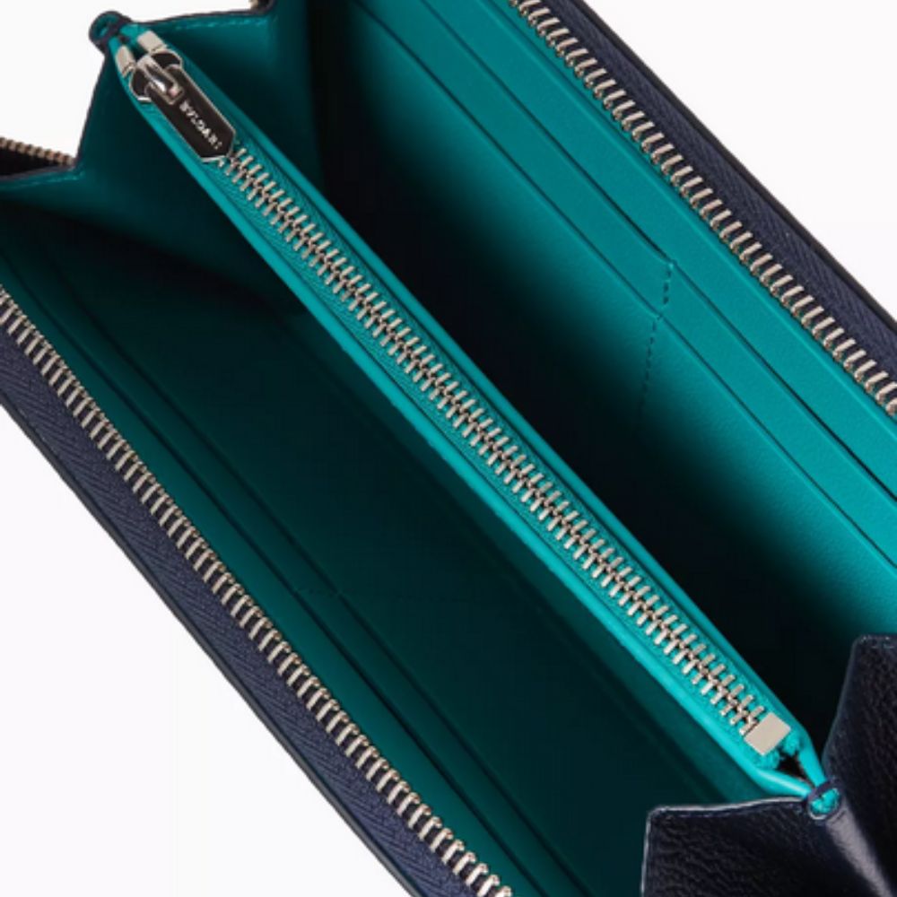 A close-up shot of the BVLGARI Zip-around Wallet in dark blue, textured leather, featuring a prominent circular silver-toned BVLGARI logo hardware with a bright teal center.