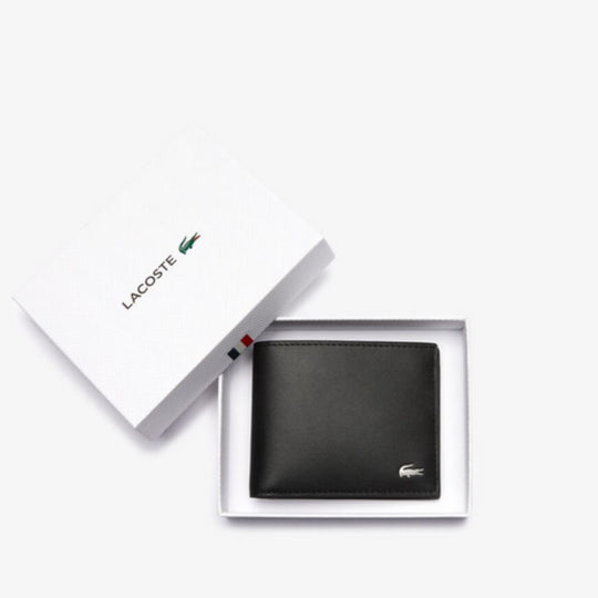 A black leather Lacoste billfold wallet with a silver crocodile logo is displayed open in a white gift box, with the lid propped open revealing the Lacoste logo and a small French flag stripe.