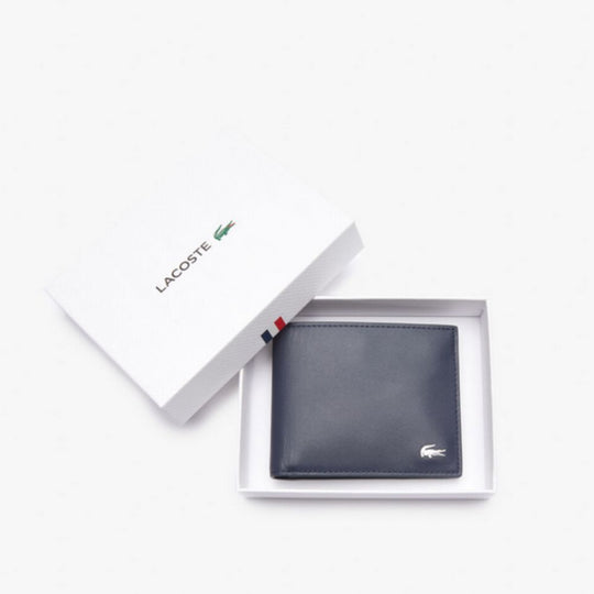 A Blue leather Lacoste billfold wallet with a silver crocodile logo is displayed open in a white gift box, with the lid propped open revealing the Lacoste logo and a small French flag stripe.