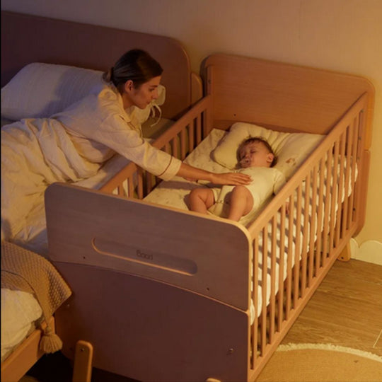 Woman tending to a child in Boori Neat 5-in-1 Convertible Cot in a warm, dimly lit room.
