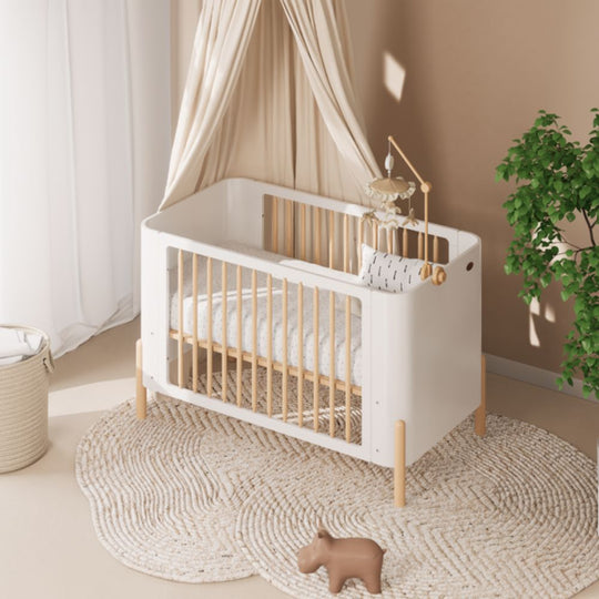 Boori Nova Cot Bed - Modern Two-Tone Toddler Convertible in Barley White & Beech in a nursery setting with a plant and rug.