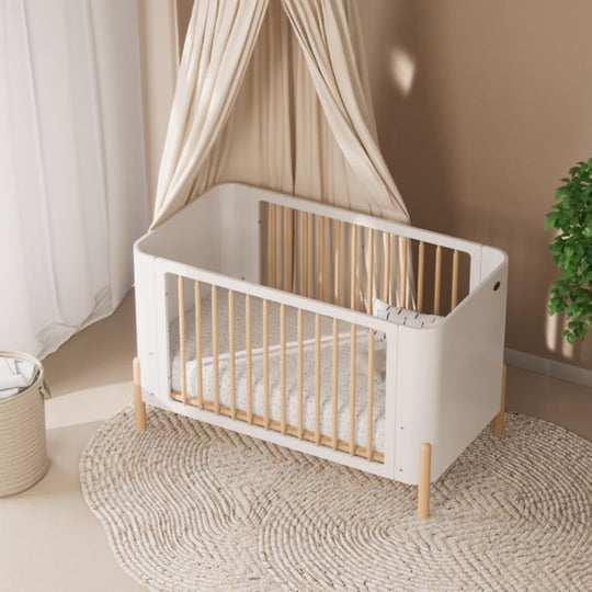 Boori Nova Cot Bed - Modern Two-Tone Toddler Convertible in Barley White & Beech in a room with beige walls and a plant.