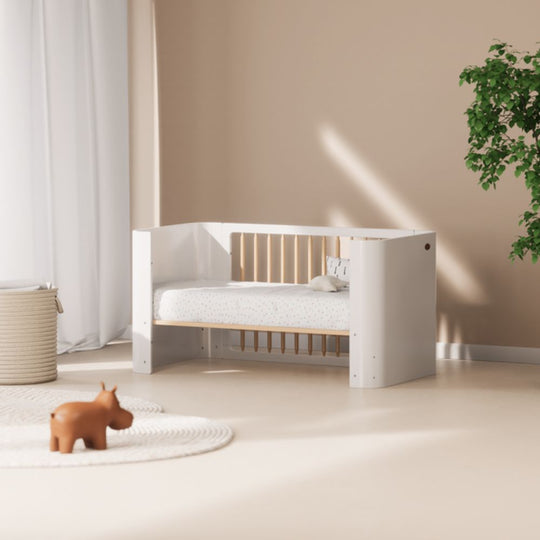 Boori Nova Cot Bed - Modern Two-Tone Toddler Convertible in Barley White & Beech in a room with a beige wall, white curtains, and a plant.