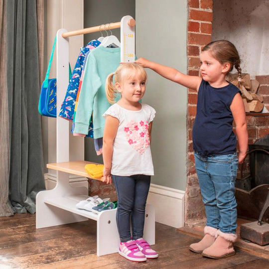 Two young girls standing next to a height chart with clothes on a rack in the background.