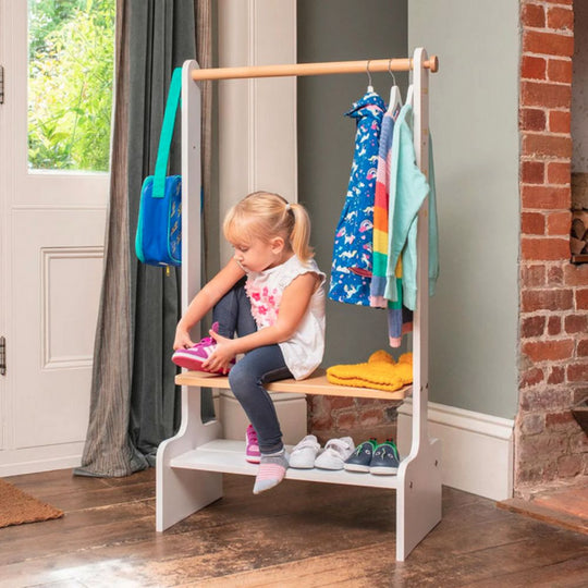 Child sitting on a small wooden stool with a clothing rack, surrounded by colorful clothes and shoes.