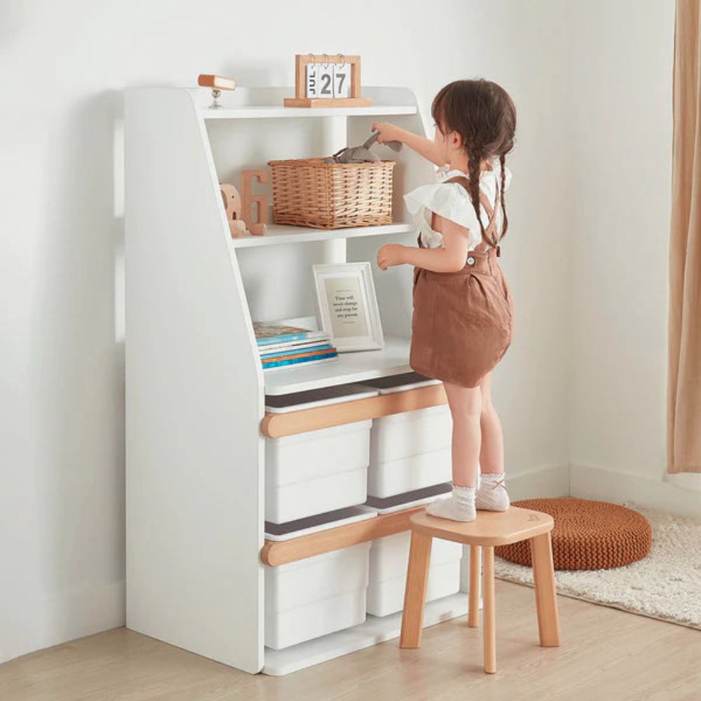 Child standing on a small wooden stool reaching into a white bookshelf.