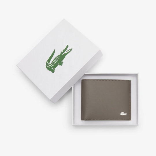 A Brown leather Lacoste billfold wallet with a silver crocodile logo is displayed open in a white gift box, with the lid propped open revealing the Lacoste logo and a small French flag stripe.