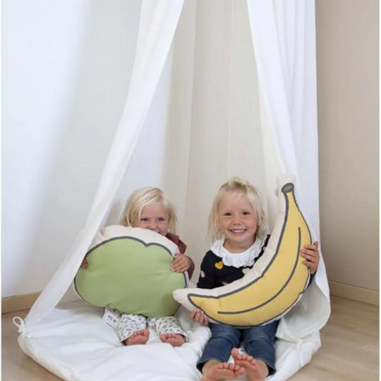 Two children sitting inside a white play tent holding pillow props shaped like broccoli and a banana.