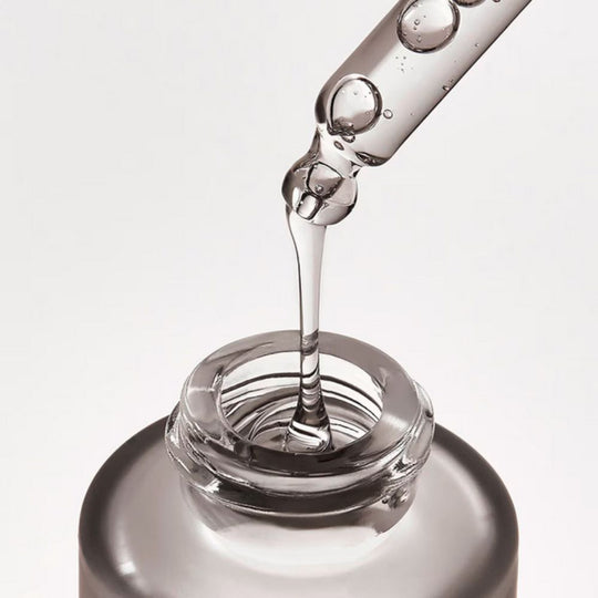 Close-up of a clear liquid serum dripping from a glass dropper into the opening of a small, translucent bottle. Bubbles are visible in the serum as it drips.