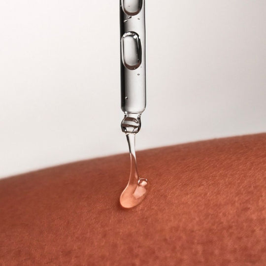 A drop of light amber or clear liquid serum is shown dripping from a glass dropper onto skin with a darker tone.