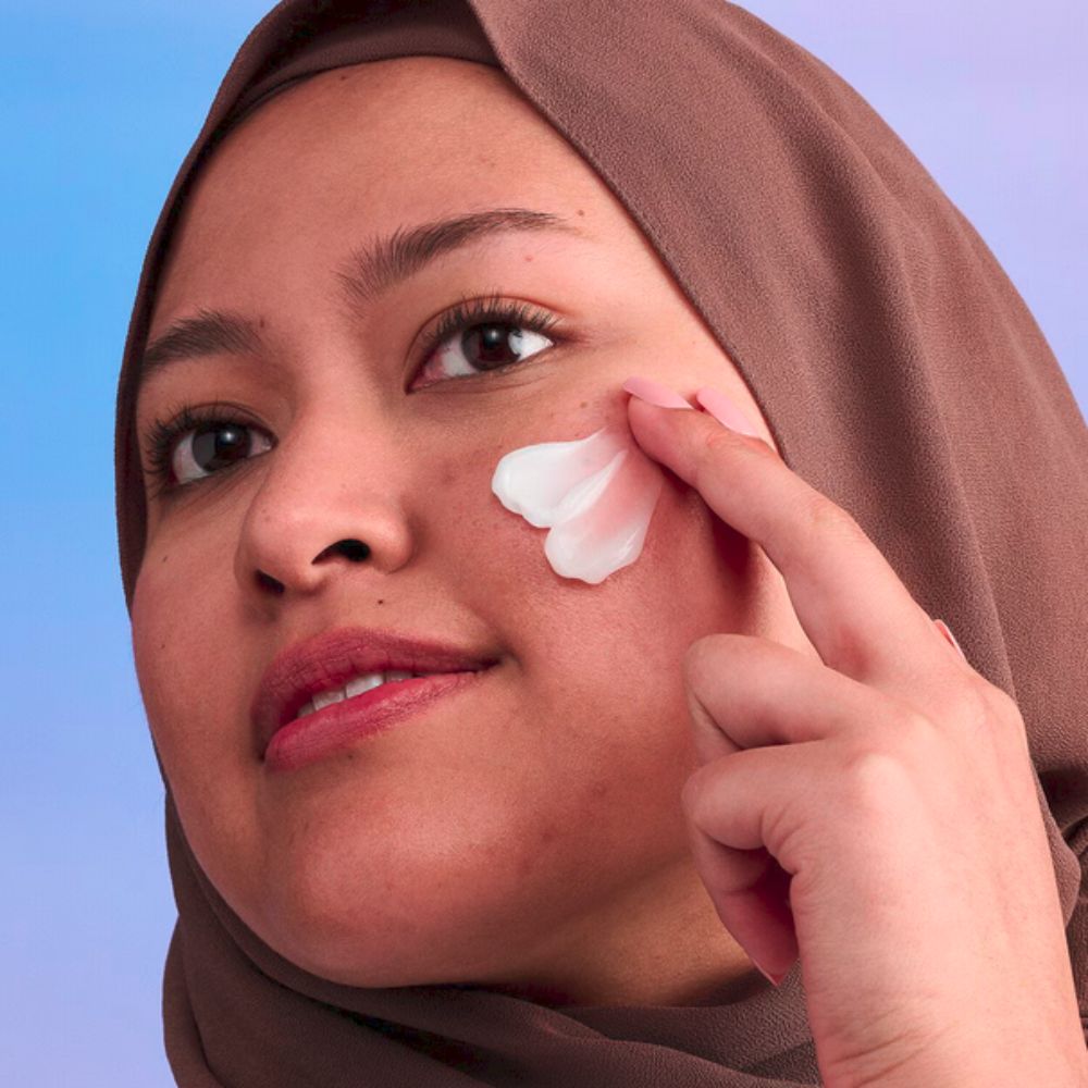 A person with a brown headscarf is applying 'DRUNK ELEPHANT face cream to their cheek.