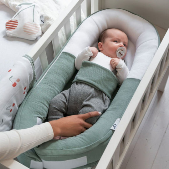 Baby in a green crib bumper with a pacifier, lying on a white crib.