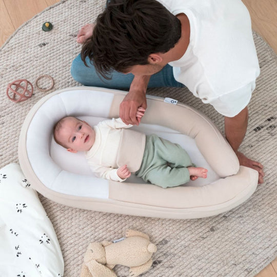 Baby lying in a white baby nest with a person interacting, on a textured rug.