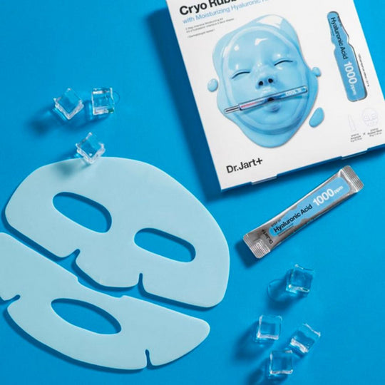 An overhead flat lay on a vibrant blue background shows a Dr. Jart+ Cryo Rubber Moisturizing Mask product box, the two-piece light blue rubber mask itself, a small ampoule of hyaluronic acid, and several clear ice cubes. The product box features an illustration of a blue face mask on the front.