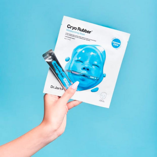 Query successful
A hand with light skin and manicured nails holds up a Dr. Jart+ Cryo Rubber Moisturizing Mask package and a tube of Hyaluronic Acid 1000 ppm ampoule against a bright blue background. The white mask package features a blue, molded rubber face mask shape and text. The silver and blue ampoule tube is labeled "Hyaluronic Acid 1000 ppm" and "Dr.Jart+".