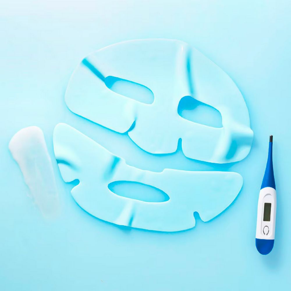 An overhead shot on a light blue surface shows the two pieces of a light blue rubber sheet mask, a white, slender pouch, and a blue and white digital thermometer. The top portion of the mask has cutouts for the eyes, and the bottom portion has a cutout for the mouth. The pouch, containing serum, is to the left of the mask. The thermometer, showing a reading, is to the right.