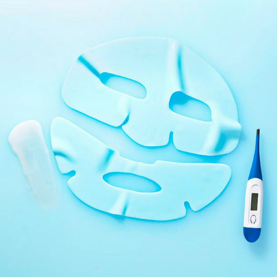 An overhead shot on a light blue surface shows the two pieces of a light blue rubber sheet mask, a white, slender pouch, and a blue and white digital thermometer. The top portion of the mask has cutouts for the eyes, and the bottom portion has a cutout for the mouth. The pouch, containing serum, is to the left of the mask. The thermometer, showing a reading, is to the right.