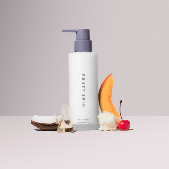 A FENTY SKIN Butta Drop Hydrating Body Milk bottle stands upright on a clean, light surface. The white bottle features "FENTY SKIN" written vertically in purple and a purple pump dispenser. Arranged around the bottle are ingredients associated with the product: a halved coconut, a small glass vial of oil, a dollop of white cream, a slice of melon, and a red cherry with its stem.