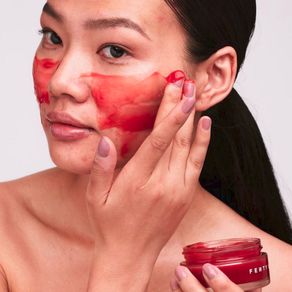 A person with dark hair pulled back applies a vibrant red FENTY SKIN Cherry Dub Blah 2 Bright 5% AHA Face Mask to their cheek with their right hand, holding the red product jar in their left. Some of the mask is already applied to their forehead and cheek, showcasing its jelly-like texture.