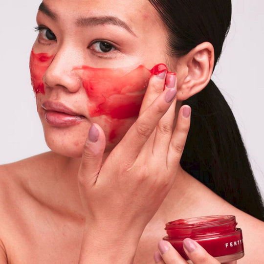 A person with dark hair pulled back applies a vibrant red FENTY SKIN Cherry Dub Blah 2 Bright 5% AHA Face Mask to their cheek with their right hand, holding the red product jar in their left. Some of the mask is already applied to their forehead and cheek, showcasing its jelly-like texture.