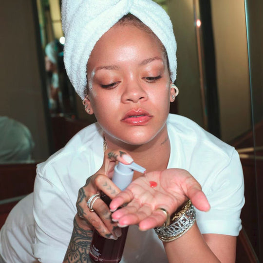 Rihanna, with a towel on her head, is holding and dispensing Fenty Skin Cherry Dub Pore Purify'r Gel Cleanser into her hand. The cherry-red cleanser is visible as she prepares to apply it.