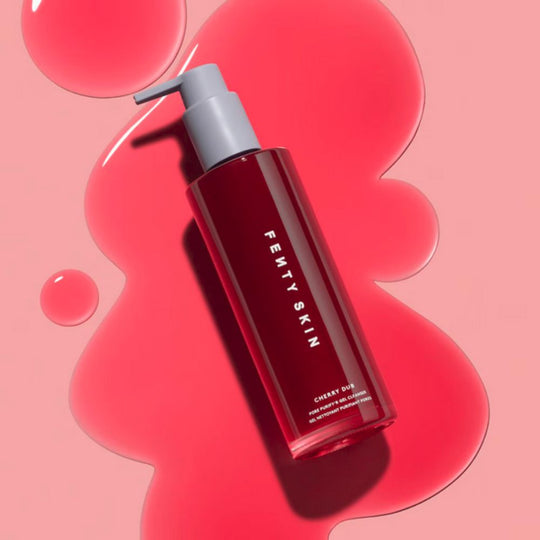 A sleek, tall bottle of Fenty Skin Cherry Dub Pore Purify'r Gel Cleanser, with a dark cherry-red product visible through its clear glass, stands on a vibrant pink and red swirled background with liquid droplets. The bottle has a light gray pump dispenser.