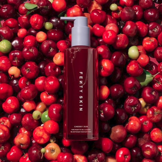 The image shows the Fenty Skin Cherry Dub Pore Purify'r Gel Cleanser, a dark cherry-red bottle with the brand name printed vertically, placed on a vibrant background of fresh cherries. The bottle has a light gray pump dispenser.