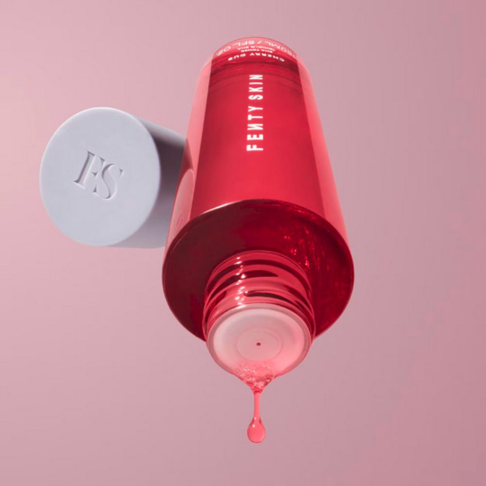 A clear, red-filled bottle of Fenty Skin Cherry Dub BHA Toner is held upside down, with a single drop of the product suspended and about to fall from its dispenser. The light gray cap of the bottle, with "FS" debossed on its top, is visible to the left, slightly out of focus.
