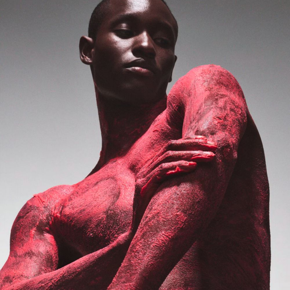A close-up shot shows a dark-skinned person with a buzz cut, seemingly applying FENTY SKIN Cherry Dub Triple Action Aha Body Scrub, evidenced by the textured, peeling reddish-pink substance covering their skin.