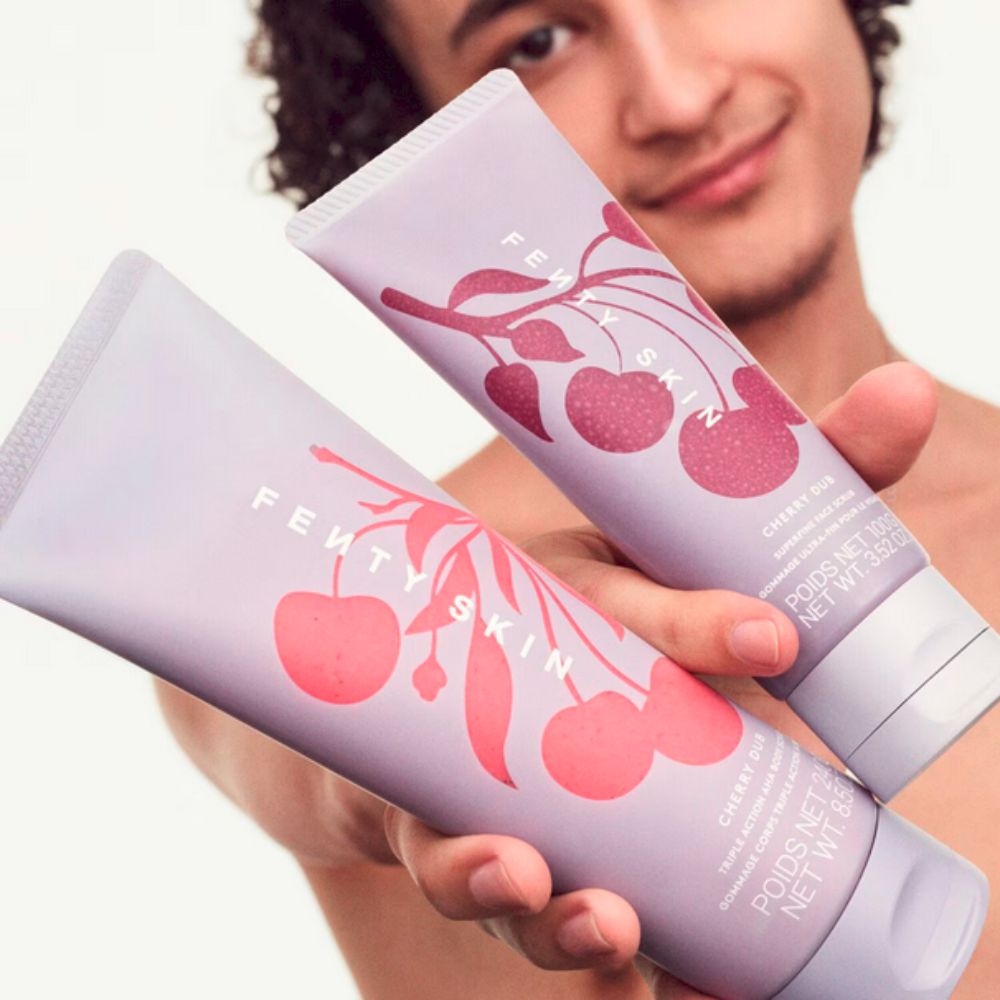 A person with curly hair holds two Fenty Skin Cherry Dub tubes towards the viewer. The foreground tube, a body scrub, features a pink cherry design on a light purple background, with "Fenty Skin" and product details visible. The background tube is a face scrub.