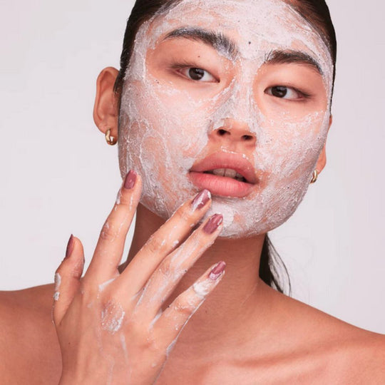 A person with dark hair pulled back applies a white, textured face mask or scrub to their face with their right hand, touching their chin. They have dark eyebrows and are looking directly at the viewer. Small gold hoop earrings are visible.