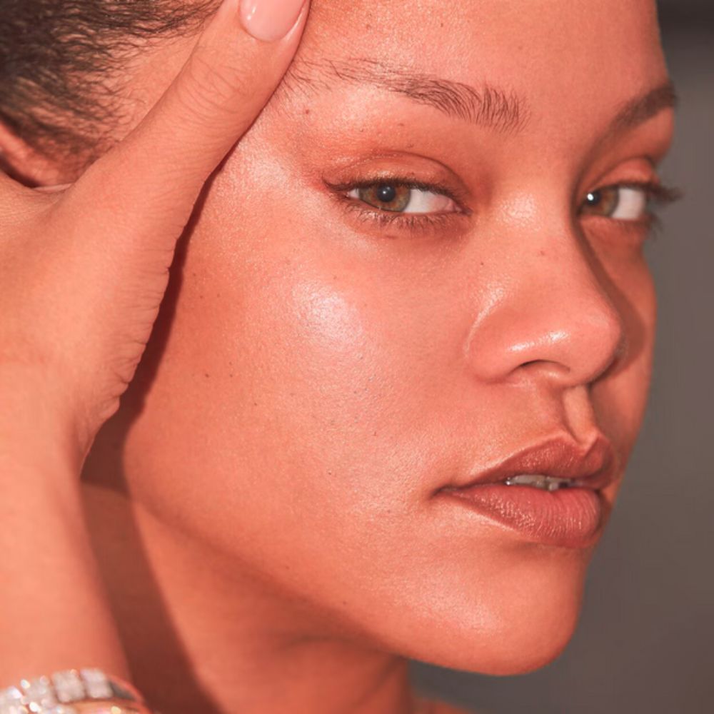 A close-up shot of Rihanna with her right hand resting on her forehead, gazing directly at the viewer. Her skin has a warm, peachy glow. She has a faint winged eyeliner and a natural lip color.