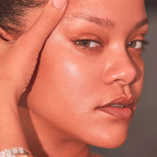 A close-up shot of Rihanna with her right hand resting on her forehead, gazing directly at the viewer. Her skin has a warm, peachy glow. She has a faint winged eyeliner and a natural lip color.