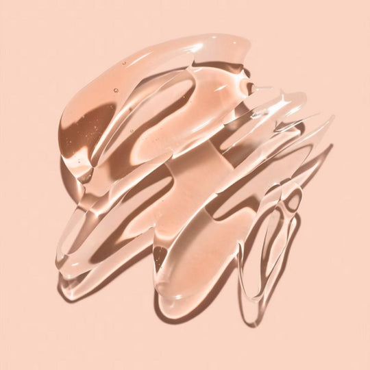 Swirls and smears of a translucent, peachy-toned liquid, likely a serum or toner, are artfully spread across a light peach background, showing the product's texture.