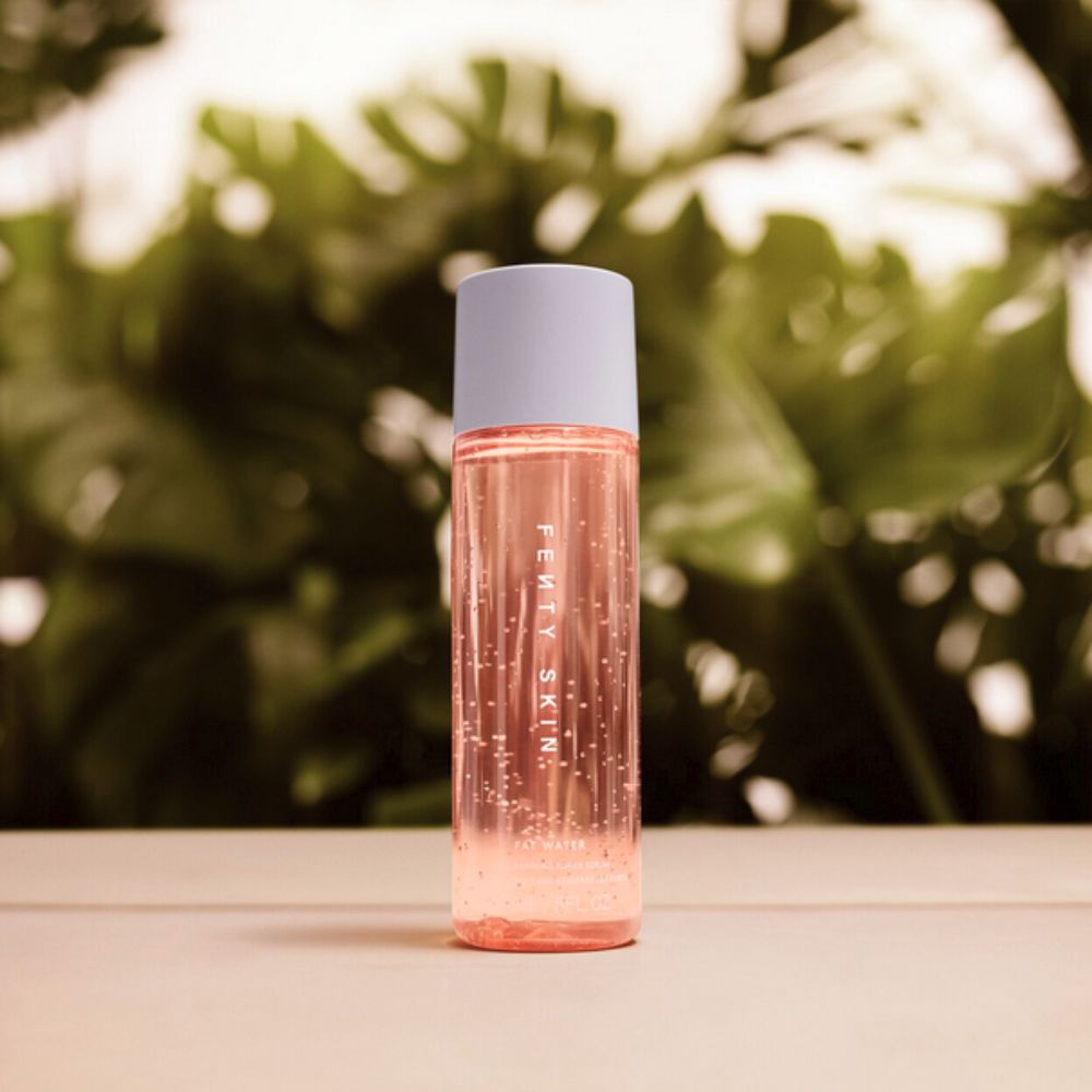 A bottle of Fenty Skin Fat Water Niacinamide Pore-Refining Toner Serum stands upright on a light surface, with a blurred background of green foliage. The clear bottle contains a pinkish liquid with small bubbles visible, and has "FENTY SKIN" printed vertically on its side. It has a light blue-grey cap.