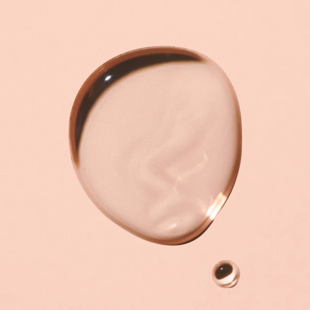 A single, large, clear droplet of a peachy-pink liquid rests on a solid, light peach background, with a much smaller, similar droplet positioned below and to the right.