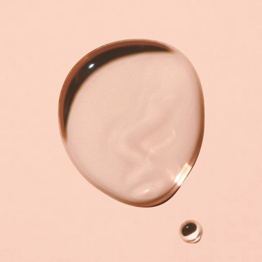 A single, large, clear droplet of a peachy-pink liquid rests on a solid, light peach background, with a much smaller, similar droplet positioned below and to the right.