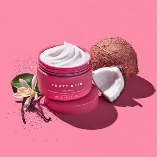 A bright pink jar of Fenty Skin "Butta Drop" whipped oil body cream sits open on a pink background, revealing a swirl of white product inside. To the right, a whole coconut and a halved coconut are visible. To the left, a vanilla bean and a vanilla flower, along with some pink glitter, complete the arrangement.
