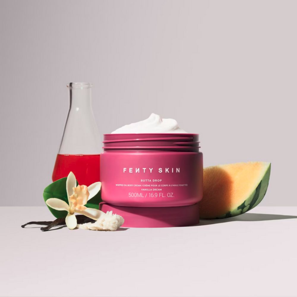 An open pink jar of Fenty Skin "Butta Drop" whipped oil body cream, showing a swirl of white product, is centered on a light surface. To its left, a glass beaker filled with a red liquid, a vanilla bean, and a vanilla flower are arranged. To the right of the jar, a slice of cantaloupe melon and a piece of coconut are visible.