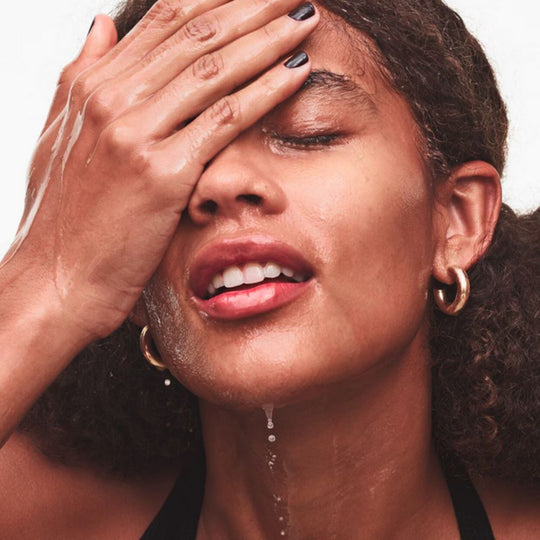 A person with curly dark hair and dark nail polish on their left hand has their eyes closed, with water dripping from their chin and forehead. Their left hand covers their eyes, and they are wearing gold hoop earrings. Their skin appears wet.