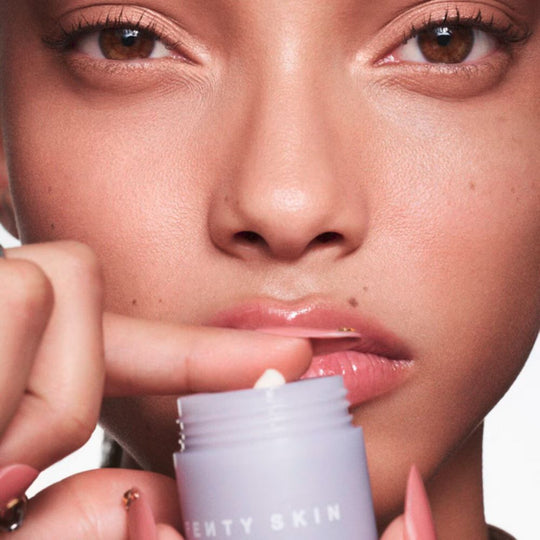 A close-up shot focuses on a person's face from the nose upwards, with a small, light purple FENTY SKIN jar held near their mouth. The person has warm brown eyes looking directly at the viewer. Their skin appears dewy and has visible freckles around the nose and upper lip area. Their lips are a glossy pink, and a small amount of white product is visible on a fingertip poised to apply. The person's nails are long and polished in a light pink shade with gold accents.