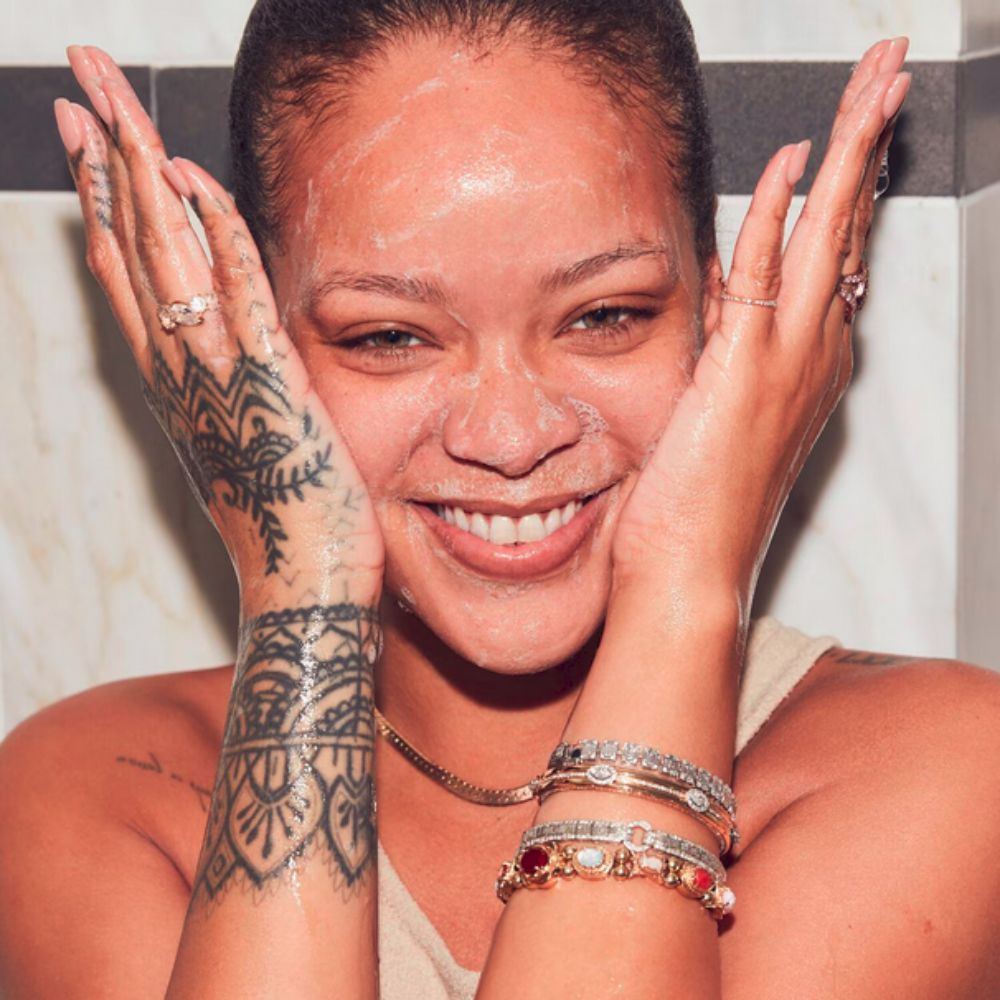 A medium close-up shot of Rihanna smiling, with a white cleanser lathered on her face. Her hands, covered in foam, are placed on either side of her face. She has several bracelets on her left wrist and rings on both hands. Her left arm features a detailed tattoo. The background appears to be a bathroom with white tiled walls.