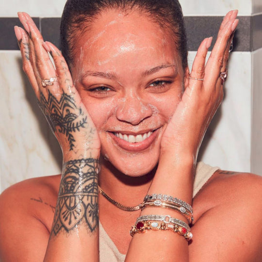 A medium close-up shot of Rihanna smiling, with a white cleanser lathered on her face. Her hands, covered in foam, are placed on either side of her face. She has several bracelets on her left wrist and rings on both hands. Her left arm features a detailed tattoo. The background appears to be a bathroom with white tiled walls.