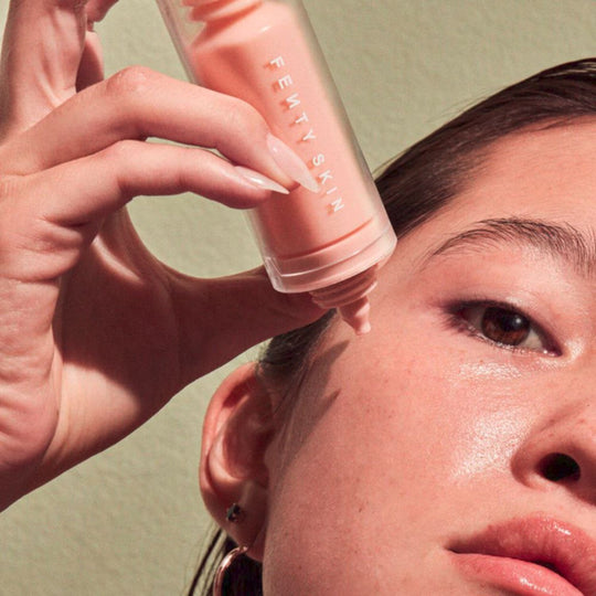 A close-up shot of a person applying a Fenty Skin product. The person's right hand, with manicured nails, holds a pinkish-orange Fenty Skin bottle, squeezing out a drop onto their cheek near their eye. This is the bottle of FENTY SKIN  Watch Ya Tone Niacinamide Dark Spot Serum. The person's skin appears dewy, and their eye is visible, looking towards the right.
