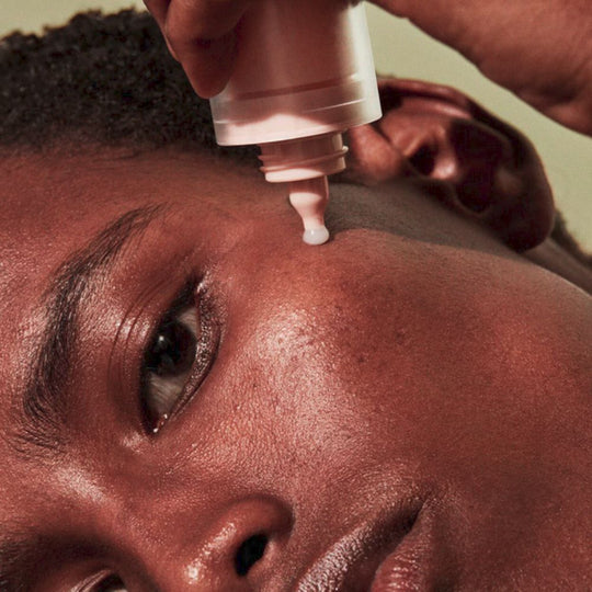 A close-up, slightly angled shot shows a person applying FENTY SKIN  Watch Ya Tone Niacinamide Dark Spot Serum to their cheek. The person has dark skin and short, dark hair. A light-colored drop of product is visible at the tip of the applicator, poised above their cheek. Their eye is visible, looking towards the right, and their skin appears well-hydrated.